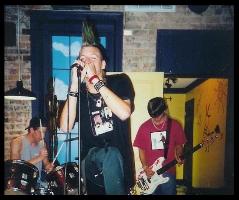 An aging punk rocker as a young man singing into a microphone at a punk rock show